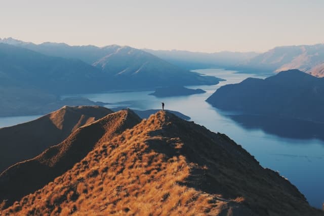 Lone figure standing atop mountain ridge overlooking dramatic New Zealand alpine landscape at sunrise